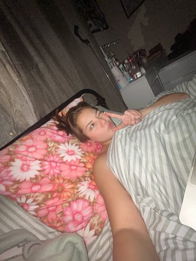 A woman with brown hair is lying in bed. She is under a striped green and white comforter. Her head is on a pillow covered in a floral pattern. She is winking and making a peace sign with her fingers near her face. The room is dark, with a cabinet and items visible in the background. The photo is taken from a slightly elevated perspective.