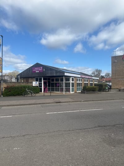 The image depicts the Thrapston Community Library in Kettering, United Kingdom. The building has a distinctive triangular facade with a sign bearing the library's name in purple. A bicycle is parked near the library entrance, which features white-framed windows and a purple door. The sky is partly cloudy. The street is paved and marked with white lines. Adjacent to the library is a building constructed with brown bricks.