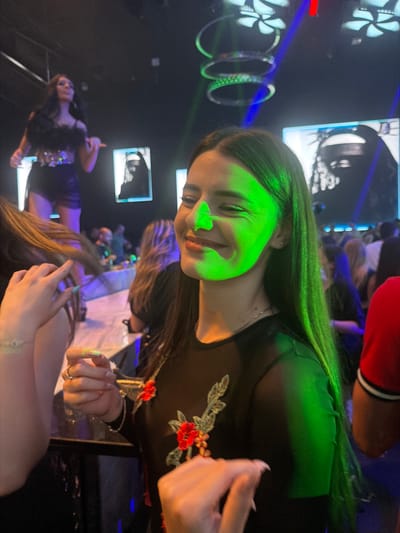 A group of young adults are socializing inside a dimly lit club. The main subject is a woman with long, dark hair, wearing a black sheer top adorned with floral embroidery. She is illuminated by green light, which casts a vibrant glow on her face and hair. Her eyes are closed, and she appears to be smiling, suggesting a moment of enjoyment. In the background, other individuals are visible, some dancing and others gathered around tables. The club's atmosphere is enhanced by colorful lighting, including blue and green beams. The environment is characteristic of a nightlife venue, with a dance floor or seating area visible. The image does not provide any specific information about the city of Plovdiv, Bulgaria, or any landmarks.