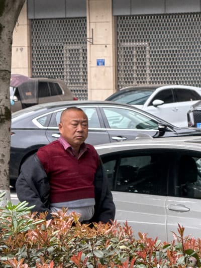 An East Asian male stands outdoors in an urban environment in Jiangjin, China. The subject, appearing middle-aged with a short haircut, wears a maroon V-neck sweater over a lighter red collared shirt, with a dark jacket draped over his shoulders. A light grey garment is visible below the sweater. He is positioned behind a low hedge of shrubs characterized by green leaves and reddish-orange new growth. In the background, multiple vehicles are parked on a paved surface, including a dark grey sedan and a silver sedan. A light-colored building facade with grid-patterned metal coverings over windows or openings is visible beyond the vehicles. A small blue sign is affixed to the building wall. A tree trunk partially obstructs the scene on the left. The man's gaze is directed slightly towards the viewer's left.