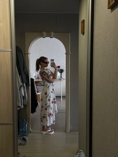 A young woman with brown hair in a ponytail, wearing sunglasses, a white sleeveless dress with red roses, and white platform sandals, takes a mirror selfie in an apartment. She has a tattoo on her left arm. In the reflection, a hallway is visible with clothes hanging on a rack, a large blue water bottle, and slippers on the floor. Further down the hallway, a round table with a vase of red flowers and a book is present, along with an arched doorway. The scene appears to be in Minsk, Belarus, based on the context provided. The lighting is dim, suggesting it's indoors during the day or evening.