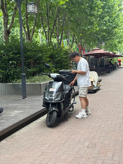 A young person in a white T-shirt and tan shorts stands on a brick-paved sidewalk next to a black motor scooter in Jiangjin, China. The person appears to be interacting with the scooter, perhaps opening a compartment. The scooter is parked on a wooden platform that runs along the edge of the sidewalk. Lush green trees and bushes line the sidewalk, and a decorative street lamp is visible to the left. In the background, outdoor seating and umbrellas suggest a cafe or restaurant.