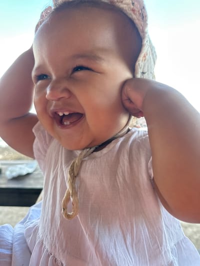 In North Kuta, Indonesia, a close-up portrait captures a young child wearing a white textured dress and a patterned bonnet. The child is seen from the chest up, with hands placed on either side of their head, pulling at the bonnet. Their mouth is open in a wide laugh, revealing small teeth. The background is softly blurred, with a hint of the sky visible above. The lighting suggests an outdoor setting, likely during the day.
