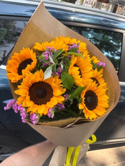 A hand holds a bouquet of sunflowers wrapped in brown paper and burlap, with a yellow ribbon tied around the base, in front of the dark teal side of a car, possibly parked on a street in Almaty, Kazakhstan. The bouquet features several large sunflowers with dark centers and bright yellow petals, interspersed with small clusters of purple flowers and green leaves. The car's window reflects the sky and trees, suggesting an outdoor setting. The hand holding the bouquet appears to be hairy and is positioned in the lower left corner of the frame. The background is slightly blurred, drawing attention to the floral arrangement and the car.