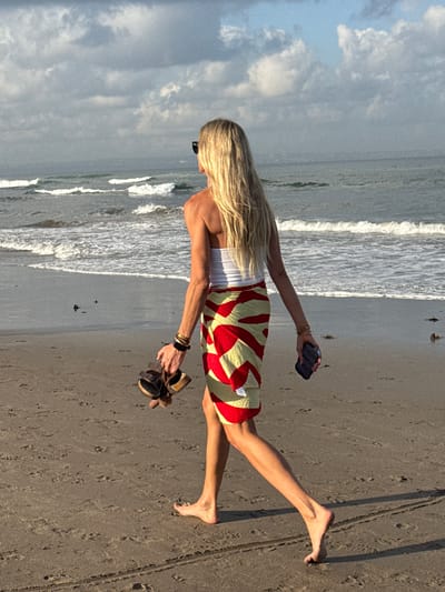 A blonde woman is walking barefoot on a sandy beach in North Kuta, Indonesia. She is wearing a white strapless top and a red and yellow patterned sarong. She is carrying a pair of sandals and a phone in her hands. The ocean is visible in the background with waves rolling in. The sky is partly cloudy.