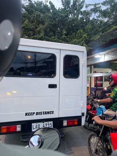 The image shows a street scene in Davao City, Philippines, at an unspecified time. In the foreground, a white vehicle with the words "KEEP DISTANCE" printed on the back is prominent. The vehicle's license plate reads "LAQ 4715". A side mirror of another vehicle is visible in the lower part of the image, reflecting the scene. To the right of the white vehicle, a person wearing a red helmet and a green and black patterned uniform is riding a motorcycle. Another person on a motorcycle is partially visible in the lower right corner. In the background, lush green trees and tangled electrical wires suggest an urban environment. A storefront with signage, including "InstaBayad" and "Instaload", is visible to the right. A poster with the image of a person and the text "I STAND WITH PRRD" is also visible on the storefront. The overall scene depicts a typical street in a bustling city, possibly during traffic.