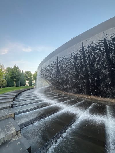 The image shows a modern, tiered waterfall structure in a park setting. The waterfall cascades down a series of dark, rectangular steps, creating a dynamic visual effect. The water is clear and appears to be flowing with considerable force. The backdrop consists of a clear blue sky with a few wispy clouds, and a line of lush green trees and manicured grass. In the distance, a person in a light-colored dress is visible standing on the steps near the cascading water. The overall scene suggests a public space designed for relaxation and aesthetic appeal. This is likely the waterfall in the "Park of the 30th Anniversary of the Victory" in Krasnodar, Russia.