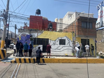 The image shows a group of people painting a mural on a building in Heroica Puebla de Zaragoza, Mexico. The building is a two-story structure with a combination of brick and concrete block construction. It has a flat roof with water tanks visible on top. The mural being painted is in progress, showing a purple flower on the left and a mountain outline on the right, on a yellow background. Several individuals are actively painting while others observe. A ladder is being used to reach higher parts of the wall. The street in front of the building has yellow and black striped markings. Tactile paving for the visually impaired is present. Overhead, there is a complex network of power lines. A sign with the word "MATAMOROS" is visible on the building. A vehicle and other buildings can be seen in the background.