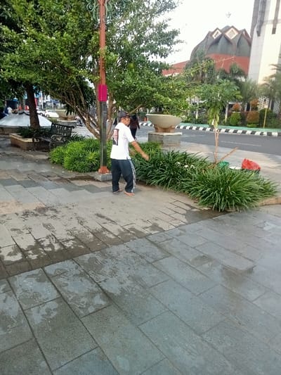 The scene takes place on a paved sidewalk in front of a large building with a dome-shaped roof. The building appears to be a mosque. The sidewalk is wet and appears to have recently been cleaned. A man in a white shirt and black pants is walking on the sidewalk. He appears to be moving his arms in a way that suggests he is stretching or exercising. There are also plants and landscaping visible. The scene is in Pati, Indonesia, at the coordinates -6.753 latitude and 111.04 longitude.