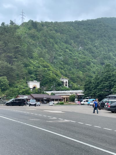 The image depicts a parking lot adjacent to a road, with a lush, densely forested mountain slope in the background. The mountain is dotted with power lines and pylons. In the middle ground, there is a low-rise building with a dark roof, possibly a cafe or restaurant, with outdoor seating and signage. Several vehicles, including black vans and cars, are parked in the lot. Two men are walking in the parking lot, one in a blue shirt and the other in a grey shirt. Further back, a white arched structure with signage is visible, and beyond that, a building with a decorative facade and a dome. The sky is overcast and cloudy. The scene appears to be located in Abaata, Abkhazia, given the presence of distinctive architecture and the mountainous landscape characteristic of the region.