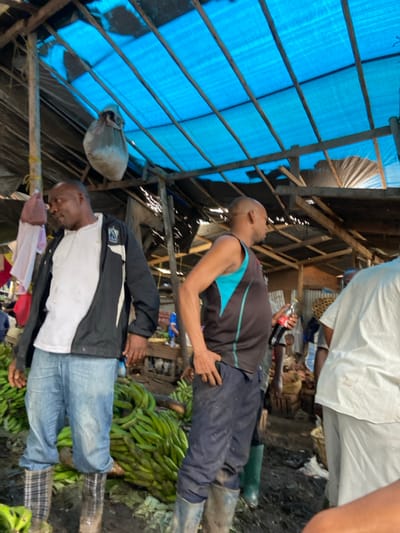 The image shows a crowded outdoor market in Dar es Salaam, Tanzania, located at the coordinates -6.801 latitude and 39.224 longitude. Three men are visible in the foreground: the man on the left is wearing a white t-shirt, a black jacket, and blue jeans. He is standing near a large pile of green bananas. The man in the center is wearing a blue and black tank top, black pants, and black boots. He is holding a bottle of Coca-Cola. The man on the right is wearing a white shirt and black pants. The market is covered with a blue tarp and wood framing. The floor of the market is dirt and there are several baskets and other items scattered throughout. There are other people present in the background but they are not visible in the image. The image was likely taken in the afternoon or evening, as the shadows are long and the light is soft.