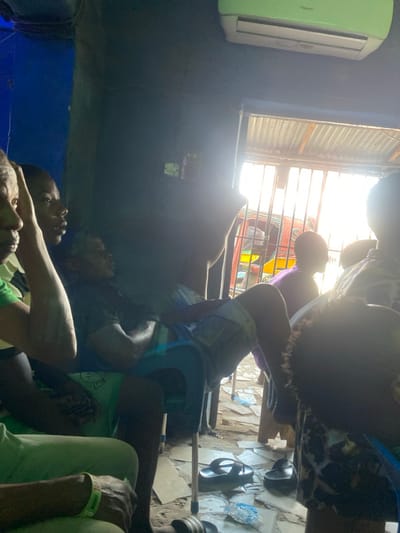 The image depicts an interior scene in Onitsha, Nigeria, likely a communal or commercial space, with bright daylight entering from an outdoor opening on the right. The room has dark blue and gray walls. A light green and white air conditioning unit is mounted high on the right wall.
Multiple individuals are present. In the left foreground, the partial profile of an older individual is visible, wearing a green garment and a light-colored wristband. Adjacent to them, a younger individual with dark skin looks towards the right. Centrally, a male individual in a dark t-shirt and light-colored patterned shorts reclines in a blue plastic chair with a metallic frame, arms crossed, facing right. Further right, a person in a purple top is partially visible, standing or leaning near the bright doorway. In the immediate right foreground, the back of a head with dark, curly hair is seen, belonging to a person wearing a patterned shirt.
The floor, appearing to be concrete or packed earth, is scattered with debris including pieces of paper, a plastic bottle, and a clear plastic sachet. A pair of black flip-flops rests on the floor near the center-right. The outdoor opening is secured by vertical metal bars, with a corrugated metal roof visible above. Through the bars, intense sunlight illuminates parts of a yellow and red vehicle, possibly a tricycle or 'Keke Napep,' and blurred outdoor elements. The overall atmosphere suggests a casual, daytime gathering.