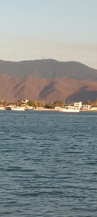 The image captures a coastal scene in Juan Griego, Venezuela. The foreground is dominated by the blue-gray, gently rippled sea. Further back, a shoreline is visible, featuring a mix of boats moored near the beach and buildings. The architecture suggests a residential or resort area. A large mountain range looms in the background, the slopes marked by shadows and topped with what appears to be a collection of transmission towers. The light suggests either early morning or late afternoon.