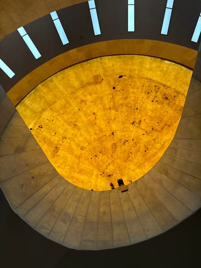 The image shows an upward view of the interior of a building, likely a dome or skylight, in Mexico City, Mexico. The central feature is a large, yellowish, translucent structure resembling a domed skylight. This element is irregularly shaped and features dark spots or markings across its surface. Two black objects are seen attached near the edge of the central element. Surrounding this is a stepped, concrete or stone structure that forms concentric rings, leading to a darker, shadowed area at the perimeter of the view. To the left side of the image, a wall with rectangular openings, possibly windows, is visible. The overall impression is an architectural view, highlighting the building's design and lighting.