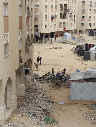 The image depicts an outdoor scene in a residential area within Khan Yunis, Palestinian Territory, characterized by extensive structural damage and makeshift living conditions. In the foreground, a multi-story, light-colored building with arched ground-level features and damaged upper-floor windows anchors the left side. Utility wires hang from this structure towards a pile of rubble consisting of concrete fragments, rebar, and shattered building materials that extends across the dusty, uneven ground.
In the mid-ground, a man in light attire is actively clearing debris with a tool. To his left, two men in dark clothing walk away from the viewer. Further right, a group of approximately four to five individuals, including adults and possibly children, are gathered around a larger accumulation of demolition debris and what appears to be the remnants of a destroyed structure or scaffolding. Several makeshift shelters, constructed from tarpaulins, fabric, and mattresses, are present, particularly on the right side of the frame, with clothes visible hanging from them and near surrounding buildings.
In the background, multiple multi-story residential buildings, similar in architectural style to the foreground structure, line a wide, unpaved area. These buildings exhibit visible damage, including shattered windows, exposed interiors, and general disarray. Clothes are hanging from many windows and balconies. The ground throughout the scene is predominantly dirt or sand, with scattered patches of sparse green vegetation and small puddles. The sky is overcast, providing diffused lighting. The overall scene suggests a recently damaged urban environment with ongoing efforts to clear debris and establish temporary shelter.