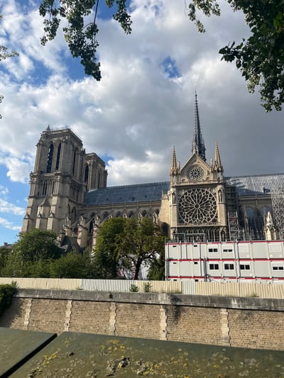 The image shows a view of Notre Dame Cathedral in Paris, France, under reconstruction. The cathedral's iconic Gothic architecture is visible, including its towers and rose windows. Scaffolding and temporary modular structures are present around parts of the building, indicating ongoing restoration work. Trees and foliage frame the scene, and a stone wall and a metal railing are in the foreground. The sky above is a mix of clouds and blue.