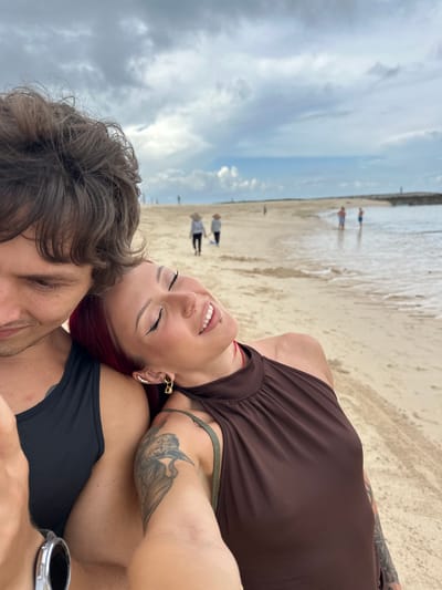 A selfie depicts a couple lying on a sandy beach, likely in Nusa Dua, Indonesia. The woman, with red hair, is wearing a brown top and has tattoos on her arm. The man is wearing a black tank top and a watch on his wrist. The beach is visible in the background with people walking along the shoreline, under a cloudy sky.