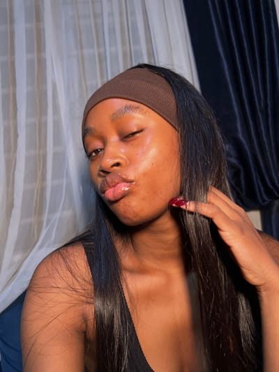 A woman with dark skin and long, dark hair is lying on a bed with one eye closed and lips pursed. She is wearing a brown headband. Her nails are painted red. One hand is resting on her hair near her cheek. She is wearing a black top. The bed has a white and grey patterned sheet, and a dark blue blanket or duvet is visible in the background.