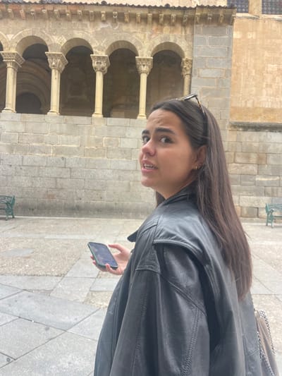 A young woman with long dark hair is standing in front of an old stone building. She is wearing a black leather jacket, sunglasses, and small gold earrings. She is holding a black smartphone in her right hand and looking back over her left shoulder. The building is an old church located in the Plaza Mayor in Segovia, Spain, with the coordinates 40.949, -4.122. The building has several arched doorways, and there is a paved courtyard in front of it. There are benches and a small tree visible in the distance.