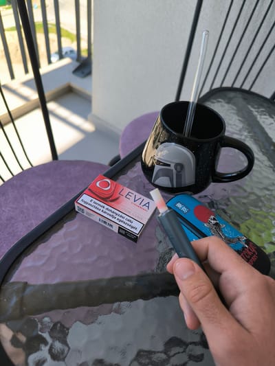 On a glass-top table, likely on a balcony in Varna, Bulgaria, a hand holds a gray vaping device. The device is partially inserted into a blue carrying case adorned with a carp design and Japanese characters. Beside these items rests a pack of LEVIA strawberry flavored tobacco sticks, featuring Hungarian text and warning labels. A black mug with a Star Wars-themed design, containing a clear glass straw, sits nearby. The table is positioned on a balcony with a railing visible in the background, overlooking a street with a parked car.
