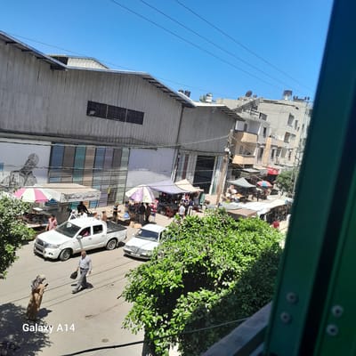 This is an outdoor, eye-level shot of a street scene in Deir el-Balah, Palestinian Territories, under a clear blue sky. The foreground is dominated by a large, vibrant green tree on the right, partially obscuring the view of the street. To the left, a woman in a tan abaya and headscarf is looking at her phone, standing on the street. An older man in a light grey button-up shirt and grey trousers is walking away from her, carrying a dark bag in his right hand.

The street is paved, with visible tire tracks and some scattered debris. A white pickup truck is parked on the left side of the street, facing away from the viewer. Further down the street, a white sedan is also parked. Several market stalls are set up along the side of the road, covered by colorful umbrellas and awnings, with people browsing and interacting. Goods are displayed on tables and racks.

In the background, there are buildings of varying heights and architectural styles. A large, corrugated metal warehouse-like structure runs along the left side of the street, featuring a large mural of a person wearing a helmet. Adjacent to it are multi-story buildings made of concrete blocks. Overhead, electrical wires are strung across the sky. The overall impression is of a busy, commercial area with a mix of residential and industrial elements.