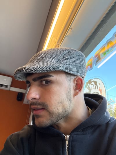 A man is posing for a photograph. He is wearing a gray newsboy cap and a black hooded sweatshirt. He has dark hair, stubble on his face, and an eyebrow piercing on his left eyebrow. The interior of a vehicle is visible in the background. A window sticker is partially visible near the top of the photograph. The location is likely Buenos Aires, Argentina, based on the presence of a window sticker of a local restaurant.