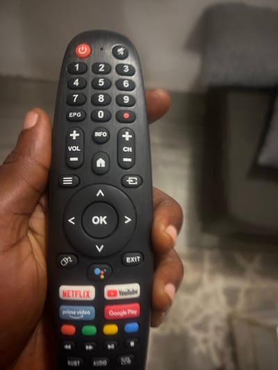 This is a close-up, indoor image depicting a person's right hand holding a black smart TV remote control. The hand, exhibiting dark brown skin, grips the remote from the left side, with the thumb positioned near the top-left edge. The remote features a red power button, mute button, a full numeric keypad (0-9), volume and channel controls, a central directional pad with an "OK" button, and dedicated shortcut buttons for Netflix (red), YouTube (red), Prime Video (blue), and Google Play (red). A Google Assistant button and media playback controls are also present. The background is out of focus, showing a dimly lit indoor setting with a partially visible light-colored, patterned tiled floor and a blurred, dark object, possibly furniture, in the upper right. The scene is located indoors within the city of Lagos, Nigeria.