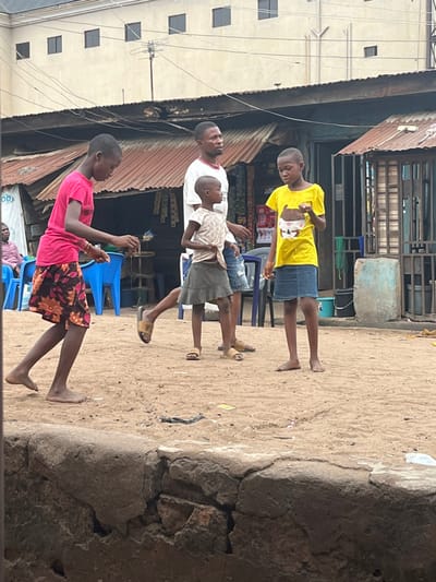 Four people stand outdoors on a sandy surface near buildings. From the front to back: a person in a pink shirt and patterned skirt, a man in a white shirt and a child in a light shirt and shorts, and another child in a yellow shirt and jean shorts. The first person is barefoot, the other three wear sandals or flip-flops. A building with a corrugated metal roof and various items visible underneath is on the left. A cement wall is on the right. The background shows other buildings and outdoor objects.