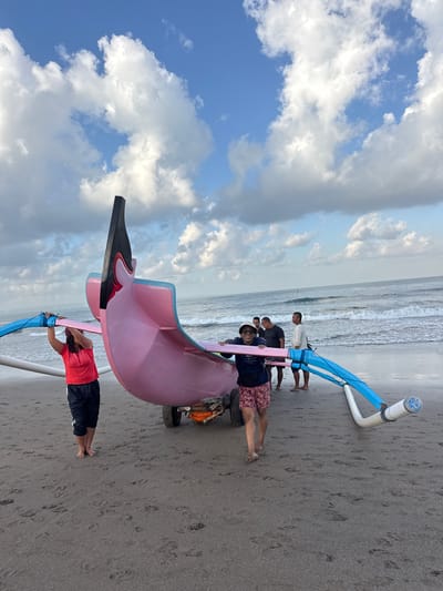 At a beach in North Kuta, Indonesia, a group of people are working together to move a colorful, outrigger canoe. The canoe is predominantly pink, with blue and white outriggers. A woman in a red shirt and black pants is on the left, holding one of the outriggers. A man in a blue shirt and patterned shorts is on the right, supporting the canoe. Behind them, three other men, dressed casually, are also involved in the effort. The scene takes place on a sandy beach with gentle waves rolling in from the ocean. The sky above is a bright blue, dotted with fluffy white clouds, suggesting it is daytime, possibly late afternoon due to the quality of light. The overall atmosphere appears to be one of cooperation and physical exertion.