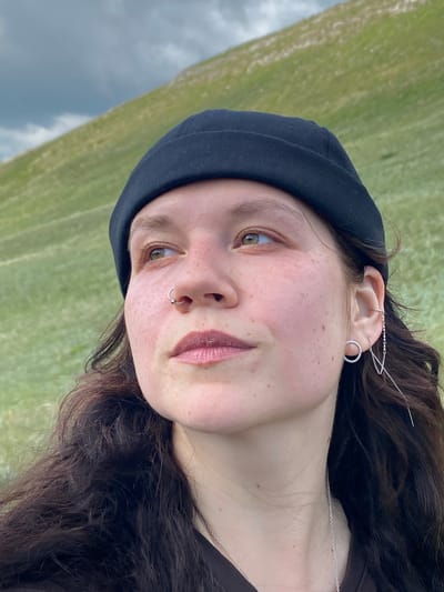 A medium shot shows a young woman with brown wavy hair, wearing a black beanie and a dark shirt, outdoors in a grassy, hilly area under a cloudy sky. She has freckles and is looking to her left. She is wearing a silver hoop earring in her right ear, and a dangling silver earring with chains and a bar in her left ear. She also has a small silver hoop piercing in her nose. The background features a green hillside and a gray sky. The location is stated as Andreevka, Russia, but this is not visually verifiable from the image alone.