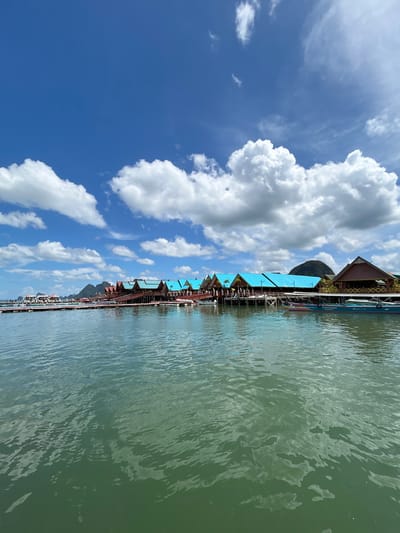 A wide-angle, eye-level outdoor shot captures the waterfront of the village of Ko Panyi, Thailand, on a clear, partly cloudy day. In the foreground, the green water of the bay is rippled by a gentle breeze, reflecting the sky above. The middle ground is dominated by a long, wooden boardwalk and stilt structures that house shops and residences. These buildings feature distinct red-tiled roofs with intricate eaves, some topped with conical spires. A series of bright blue roofs catch the eye, creating a vibrant contrast against the natural surroundings. Several longtail boats, common to this region, are moored along the boardwalk, some with passengers disembarking or preparing to depart. A few individuals are visible on the boardwalk, adding a human element to the scene. In the background, lush green, mountainous islands rise from the horizon, silhouetted against a bright blue sky dotted with fluffy white cumulus clouds. The overall impression is one of a bustling, picturesque floating village in a tropical setting.