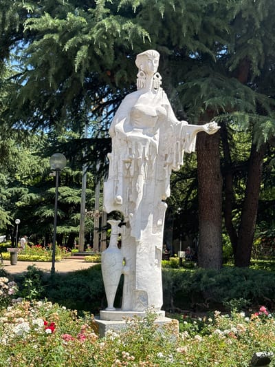 This image depicts a large, white, abstract sculpture of a woman standing in a park. The sculpture is situated in front of a backdrop of lush green trees, with a clear blue sky visible above. In the foreground, a dense bed of flowering roses, in shades of white, pink, and red, surrounds the base of the sculpture. A lamppost is visible to the left of the sculpture, and in the background, there are other park features such as benches, pathways, and more trees. A few people are present in the background, scattered throughout the park. The sculpture itself is characterized by its textured surface and draped clothing, with large, intricate earrings adorning the woman's head. Her right arm is extended, holding a small object, possibly a flower or a piece of fruit. The sculpture is located in the Rose Garden of Kazanlak, Bulgaria.