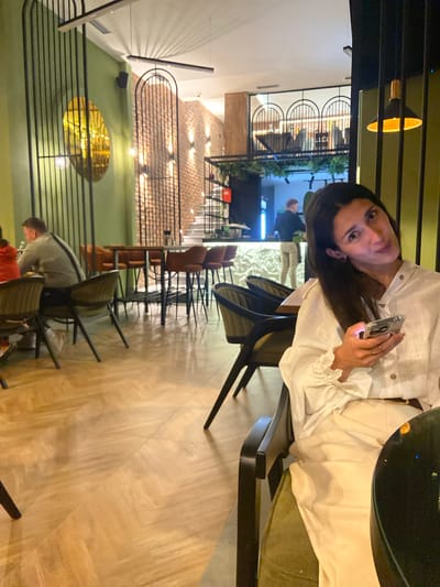 This media file captures an indoor scene, likely a restaurant or cafe in Sutomore, Montenegro, during the evening or night, given the reliance on artificial lighting.

The primary subject is a woman seated at a dark table on the left side of the frame. She has long dark hair and is wearing a white collared shirt with distinctive ruffled cuffs. She is holding a silver-colored smartphone in her right hand and is looking directly at the camera with a neutral to slightly smiling expression. She appears to be sitting on a dark green upholstered chair.

The background reveals a multi-level interior with a modern design. The floor is covered in light-colored wooden parquet. In the midground, numerous dark-framed chairs with dark upholstery are visible, arranged around tables. A prominent feature of the decor is a series of black metal railings, some with arched top sections, separating different areas. Behind these railings, a wall with a warm, textured brick-like finish is illuminated by several small lights.

Further into the background, another individual, a man, is seen bending over near other tables and chairs. Sections of green wall and some artificial greenery add to the ambiance. A large, round golden light fixture or decorative element is visible lower in the background, reflecting light. Overhead, various modern light fixtures cast a warm glow. The overall atmosphere is chic and inviting. No discernible text is visible in the image.