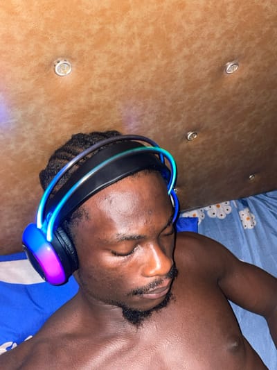 A topless, dark-skinned man is lying on his side on a bed. His eyes are closed, and he appears to be sleeping. He has short, dark hair and a beard. He is wearing headphones with blue and pink lights. The bed has a brown headboard with decorative buttons and blue floral-patterned sheets. The image is taken from a top-down perspective.