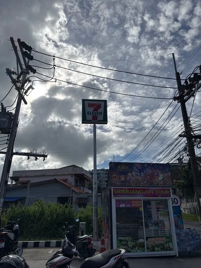 In Rawai, Thailand, under a heavily clouded sky with sunlight breaking through, a 7-Eleven sign is prominently displayed on a tall pole. Below the sign, a small business called "SRIMUANG ON TOUR" operates from a kiosk, offering tourist information, internet, and SIM cards. The kiosk features images of elephants and promotional material. To the left, a transformer and utility poles with numerous wires are visible, indicating urban infrastructure. The foreground shows several parked motorcycles, suggesting a common mode of transportation in the area. A low building with dense vegetation in front of it is also part of the scene, hinting at residential or commercial properties adjacent to the street. A speed limit sign of 50 is visible on the right.