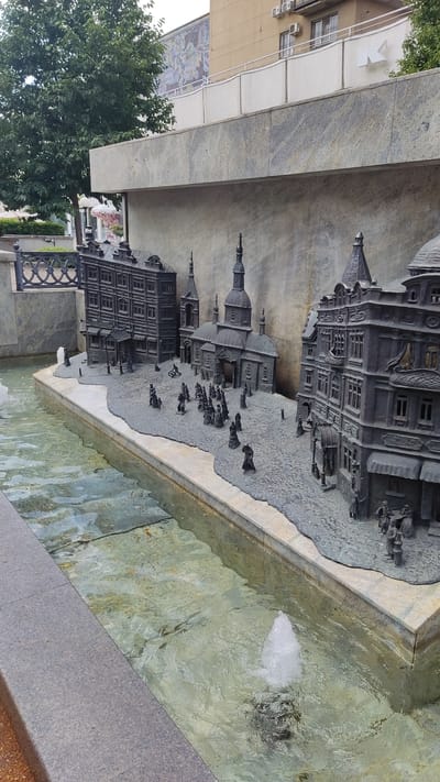 The media file captures a close-up, rotated view of a miniature diorama depicting historical architecture and figures, likely situated in Krasnodar, Russia. The diorama is integrated into a fountain, with clear, shallow water flowing around its base, suggesting it's an outdoor public installation.

The diorama itself features intricate, dark metal models of buildings resembling historical Russian architecture, complete with towers, domes, and detailed facades. Small figures, also in dark metal, are scattered throughout the scene, appearing to represent people or animals engaged in various activities. Some figures are in carriages, while others seem to be walking. The diorama is mounted on a textured, grey surface that mimics cobblestones or a similar ground texture. The base of the diorama and the fountain's edge are made of polished, marbled stone.

In the background, beyond the diorama, a building with a light-colored facade and visible windows is present, along with air conditioning units mounted on its side. A large, leafy green tree is also visible on the left side, partially obscuring the background building. The water in the fountain is clear with a slight greenish tint and shows ripples and a small jet of water. The lighting suggests it is daytime, likely with overcast or diffused sunlight, as there are no harsh shadows.

No discernible text is visible within the diorama or its immediate surroundings. The overall impression is a piece of public art or a historical monument, designed to evoke a sense of past times.