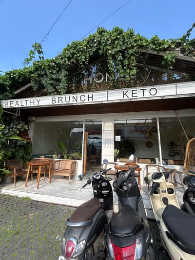 The image shows the exterior of a cafe in North Kuta, Indonesia, identified by the text "HEALTHY BRUNCH KETO" on the facade. The cafe has a modern design with large glass windows and a wooden frame covered in lush green vines. In front of the cafe, there are several parked motorcycles. Rattan furniture, including chairs and a bench, is arranged on a wooden deck. The ground in front of the deck is a permeable pavement with grass growing in the gaps. The sky is clear and blue. There are no people clearly visible in the immediate foreground or interacting with the cafe.