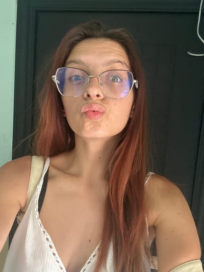 A young woman with reddish-brown hair and fair skin is shown from the shoulders up, wearing large, silver-rimmed glasses and a white, textured tank top. She is making a kissing face, with her lips puckered. Her hair is slightly messy and falls over her shoulders. A strap of a cream-colored bag is visible over her left shoulder. She appears to be indoors, in front of a dark doorway or wall. The lighting suggests it is daytime, possibly near a window. The image does not contain any identifiable landmarks or elements that would place it in the city of Tryavna, Bulgaria.