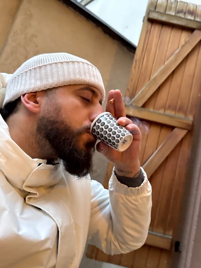 The image displays an adult male in an outdoor setting in Strasbourg, France. The subject, seen in profile, wears a light-colored ribbed knit beanie and a white hooded jacket. He has a dark, full beard, and his eyes are closed. He is actively drinking from a small disposable cup held by his right hand; the cup is white with a repeating black and grey circular pattern and a small black graphic near its base. Simultaneously, a lit cigarette is held between two fingers of the same hand, positioned above the cup, with a visible plume of smoke rising. A tattoo with text, appearing to spell "VELMIA" or similar, is visible on his left wrist/forearm. The background to the left consists of a plain, light-brown wall, while to the right, a light-colored wooden door or gate with vertical planks and diagonal support beams is present. A light, indeterminate overhead structure or sky is visible at the top of the frame.
