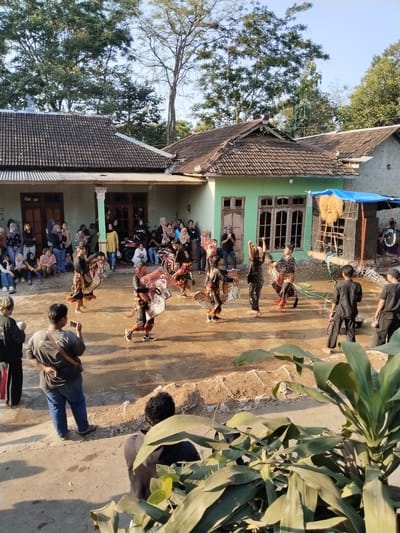 In Kerjo, Indonesia, a cultural performance is taking place outdoors in front of traditional houses. Several individuals, dressed in dark attire and ornate headdresses resembling horses, are engaged in a dance. They are surrounded by a crowd of spectators, many of whom are seated and watching the performance. The scene is set against a backdrop of houses with tiled roofs and lush green trees. A sound system with speakers and what appears to be a prop are visible on the right side of the image. The ground is dirt and sand.