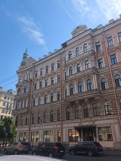 The image shows a wide, ornate, pink building in Saint Petersburg, Russia, on a clear, sunny day. The building has multiple floors with numerous arched windows, decorative molding, and balconies. A small green turret with a decorative spire tops one corner of the building.
In front of the building, a street scene unfolds. Several cars are parked or driving on the road. On the sidewalk, there are pedestrian crossing signs and traffic lights, indicating a busy urban environment. A tree with green foliage stands near the crossing. The store at the ground level of the building has a sign that reads "MANDERS" and below that, in Russian, "краски обои плитка паркет ковры карнизы - дизайн" (paints, wallpaper, tiles, parquet, carpets, moldings - design). The sky above is a bright blue with a few wispy clouds and some overhead wires, suggesting a typical summer day in the city. The architecture of the building and the presence of the Russian text on the storefront confirm the location as Saint Petersburg.