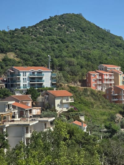 The image depicts a hillside residential area in Prijevorac, Montenegro, under a clear blue sky. The foreground features lush green trees and foliage, with the middle ground dominated by several multi-story buildings constructed on the sloping terrain. These buildings are a mix of architectural styles, featuring red tile roofs, white and light-colored facades, and balconies with glass railings. Some buildings appear to be apartments or villas. The background shows a densely vegetated green hill, characteristic of the Montenegrin landscape. There are no people visible in the image. The overall scene conveys a sense of a sunny day in a coastal or mountainous region.