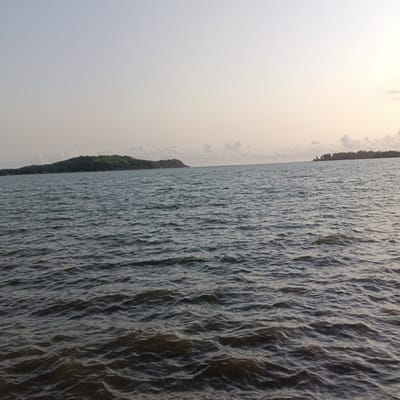 The image shows a body of water, likely a river or bay, under a pale sky. Two landmasses, possibly islands, are visible in the distance. The larger one on the left is covered in trees, while the smaller one on the right has sparse vegetation. The water surface is rippled, indicating a slight breeze. Given the prompt's location, this could be near Badem, India, specifically the Terekhol River estuary. No people, subjects, objects, actions, or interactions are directly visible in the scene.