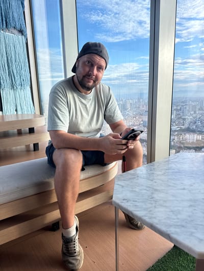 A middle-aged Caucasian man with fair skin and a beard is sitting on a light-colored bench in front of a large window overlooking a city. He is wearing a light gray t-shirt, dark blue shorts, a black baseball cap, white socks, and gray sneakers. He is holding a black smartphone with a red lanyard in his hands and looking at the camera. Behind him, a large blue tapestry with fringe hangs on the wall. To his left, a wooden table with two levels is visible. The window behind him offers a panoramic view of a dense city with numerous buildings, a hazy sky with clouds, and distant mountains. The scene appears to be set in a modern building, possibly a hotel or a restaurant, in the city of Hà Nội, Vietnam.