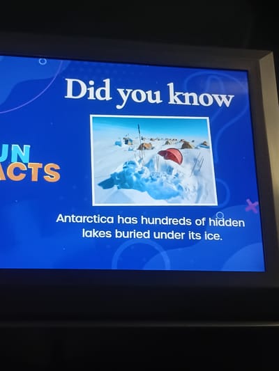 The image is of a digital screen displaying a fun fact about Antarctica. The screen is framed by a dark border, suggesting it is likely a monitor or display within an establishment. On the screen, in large white lettering, it says "Did you know". To the left, partially visible, are the words "UN FACTS" in large, bold, orange letters. In the center of the screen is a photograph of a snowy landscape with several tents and a red umbrella sticking out of the snow. Below the image, in white text, it reads: "Antarctica has hundreds of hidden lakes buried under its ice." The background of the screen is a vibrant blue with faint white circular and wave-like graphics. The image does not provide any information to suggest a location in Dubai, United Arab Emirates.