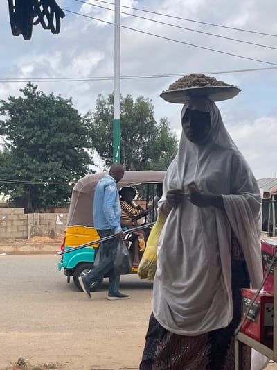 In Bukuru, Nigeria, a woman in a white hijab balances a large tray of brown, round items on her head. She is wearing a patterned skirt and appears to be holding a mobile phone in her hands. To her left, a man in a light blue collared shirt and dark pants walks with two long metal poles and a black plastic bag. Behind him, a yellow and blue auto-rickshaw, also known as a Keke Napep, is parked. Inside the Keke Napep, a person wearing a striped shirt is visible. The scene is outdoors, with a cloudy sky overhead and overhead power lines. Trees and a low brick wall are visible in the background, along with some market stalls to the right.