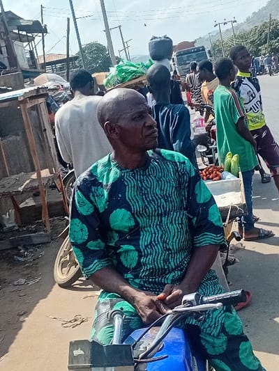 A street scene reveals a man on a parked motorbike in the foreground, wearing a patterned green and dark blue top. He appears to be adjusting something on the motorbike. In the background, people walk along a road with a truck visible further back. The setting seems to be outside during daytime. Wooden structures and buildings line the street.