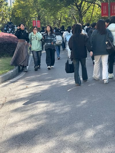 The image depicts an outdoor scene on an asphalt walkway within what appears to be a university campus or public park in Jiangjin, China, under bright daylight. A group of approximately five young individuals, primarily females, walk from left to right and towards the background on a grey asphalt path.

On the far left, a female with dark hair wears a black long-sleeved top and a long plaid brown and grey skirt, with dark boots visible. She holds a small dark object in her right hand and looks towards the left. Next to her, a female with dark hair wears a light green hooded sweatshirt with "SNJ" visible on the chest, dark jeans, and dark sneakers. She has her right hand near her face. Adjacent to her, a female with dark hair wears a blue, white, and grey patterned long-sleeved top, dark jeans, and white sneakers, holding a dark smartphone in her right hand.

Further into the frame, a female with dark long hair, seen from behind, wears a black jacket and dark flared jeans, carrying a black shoulder bag on her left side. To her right, another female, also seen from behind, wears a dark denim jacket, light-colored flared pants (possibly white or beige), and light-colored shoes.

In the background, a larger crowd of individuals, many carrying backpacks, is visible on the walkway and a sloped grassy area to the left. The environment includes a concrete curb separating the asphalt path from a green grassy area and low reddish-brown shrubs on the left. Numerous green-leafed trees are present, particularly in the background, with two red banners displaying white Chinese characters hanging vertically among them. Long, diagonal shadows are cast across the asphalt path by unseen elements, suggesting the sun is at an angle. No explicit interactions are observed between the individuals, who appear to be walking along the path.