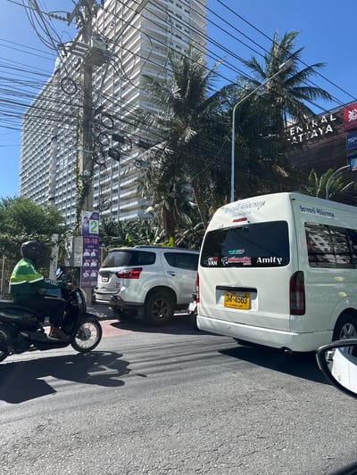 In Pattaya City, Thailand, on a sunny day, a street scene unfolds with traffic and city infrastructure. A white minivan is prominently featured in the foreground, displaying a Thai license plate that reads "34-4563". Various stickers adorn the rear window of the minivan, including one that says "Amity". To the left of the minivan, a white SUV is visible. In the background, a tall, modern white building with many balconies dominates the skyline, alongside lush green palm trees and a utility pole heavily laden with tangled electrical wires. A person wearing a green and white jacket and a black helmet is riding a black scooter on the left side of the image, casting a shadow on the asphalt road. Posters are attached to the utility pole, one with a picture of a person and the number '2', and others with multiple portraits and Thai text. The road surface shows signs of wear with cracks and patches.