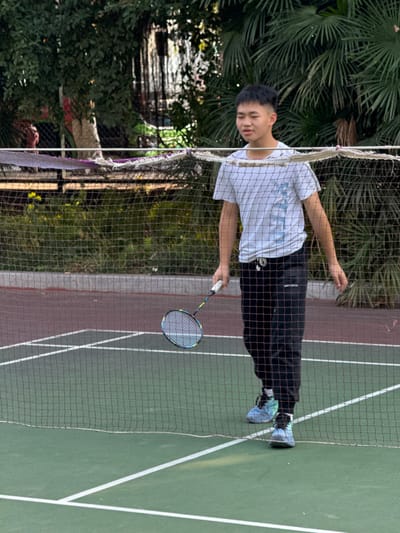 An outdoor scene in Jiangjin, China, depicts a single East Asian male standing on a green sports court, bisected by a badminton net. The individual, appearing to be in his late teens or early twenties, wears a white t-shirt with light blue markings, black athletic jogger pants, and blue/teal high-top athletic shoes. He holds a black, blue, and yellow badminton racket in his right hand, angled downwards. The badminton net, exhibiting frayed edges, stretches horizontally across the mid-ground. The court features white boundary lines on a green surface, with a section of reddish-brown court visible beyond the net. The background consists of dense green foliage, including palm fronds and other trees, under what appears to be natural lighting. No other individuals or direct interactions are visible.